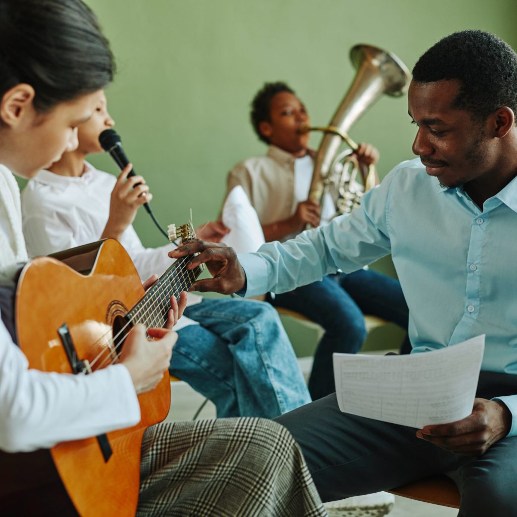 Young teacher of music consulting Asian schoolgirl with acoustic guitar at lesson while both sitting against boys with trumpet and microphone